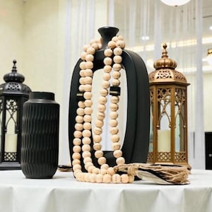 May include: A decorative display featuring a black vase with a unique shape, draped with a strand of light-colored wooden beads. A gold lantern and a black lantern are also present, all set on a white tablecloth.