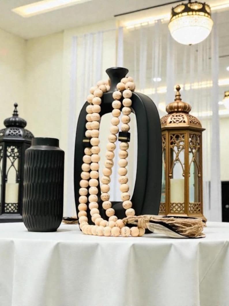May include: A decorative display featuring a black vase with a unique shape, draped with a strand of light-colored wooden beads. A gold lantern and a black lantern are also present, all set on a white tablecloth.