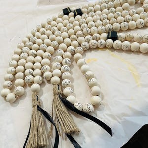 Puede incluir: Tres rosarios de madera blanca con borlas negras y detalles negros. Cada cuenta est&aacute; inscrita con caligraf&iacute;a &aacute;rabe.