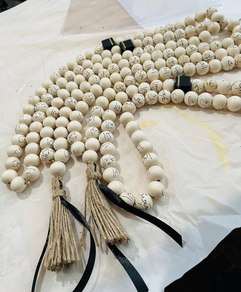 Puede incluir: Tres rosarios de madera blanca con borlas negras y detalles negros. Cada cuenta est&aacute; inscrita con caligraf&iacute;a &aacute;rabe.