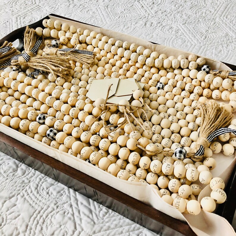 May include: A wooden tray filled with numerous light-colored wooden beads, each inscribed with names. The beads are arranged in rows and adorned with jute tassels and black and white checkered accents. A few blank tags are also present.