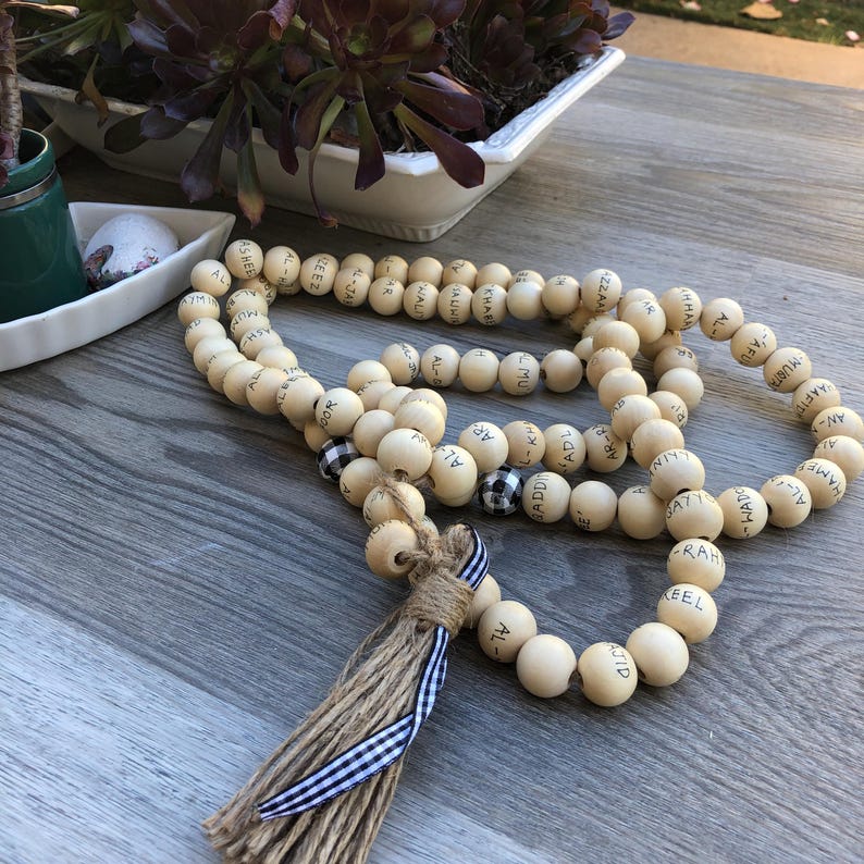 May include: A wooden bead garland with black lettering and a jute tassel. The beads are light-colored with black lettering. Some beads have a black and white checkered pattern. The tassel is tied with a blue and white striped ribbon.