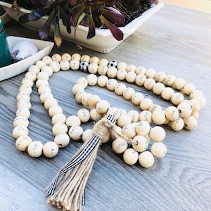 May include: A cream-colored beaded garland with black lettering. The garland features a tassel with a striped ribbon and twine. The beads are arranged in a long, flowing pattern, suitable for home decor.