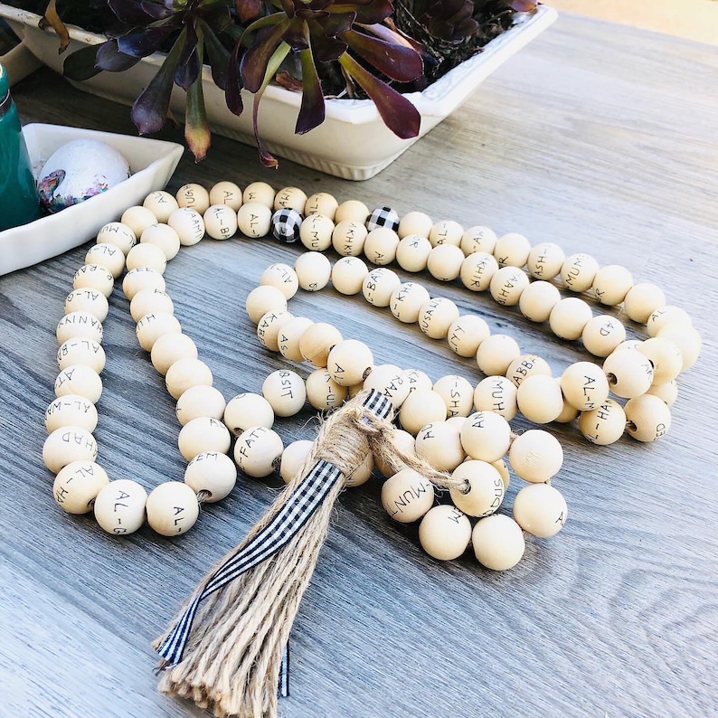 May include: A cream-colored beaded garland with black lettering. The garland features a tassel with a striped ribbon and twine. The beads are arranged in a long, flowing pattern, suitable for home decor.