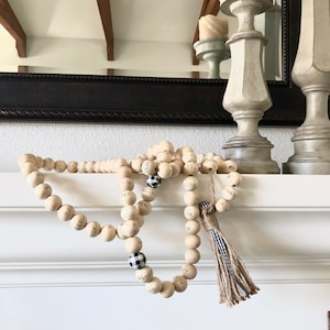May include: Decorative wooden bead garland with printed Arabic script and a tassel. The garland features natural wood beads, interspersed with black and white checkered beads. The tassel is made of twine and striped fabric.
