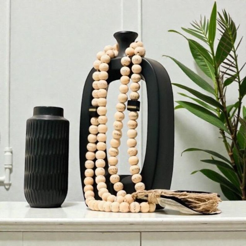 May include: A black ceramic vase with vertical ridges stands next to a black, open-frame decorative piece. A strand of light-colored wooden beads is draped over the decorative piece. A bundle of twine and feathers rests on the surface.