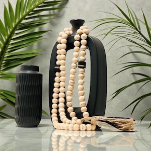 May include: A black ceramic vase with a textured surface and a tall, black, open-frame decorative piece. A long strand of light-colored wooden beads is draped over the decorative piece. Green palm fronds are in the background.