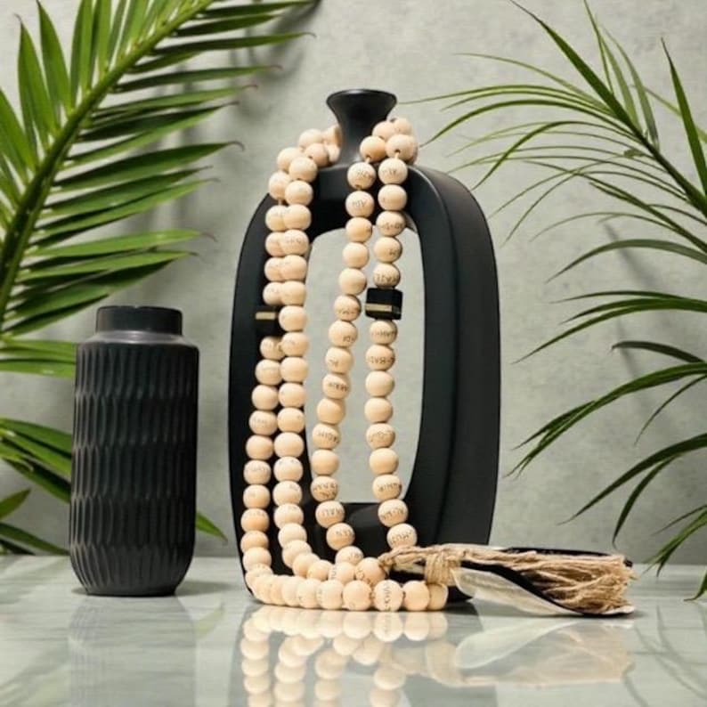 May include: A black ceramic vase with a textured surface and a tall, black, open-frame decorative piece. A long strand of light-colored wooden beads is draped over the decorative piece. Green palm fronds are in the background.