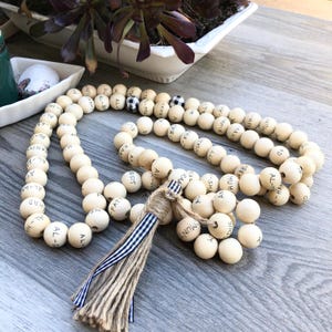 May include: A cream-colored wooden bead garland with black lettering and a jute tassel. The beads are strung together, with a few featuring a black and white checkered pattern. The tassel is adorned with a blue and white striped ribbon.