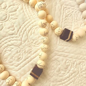 Puede incluir: Un collar de cuentas de oraci&oacute;n blancas con cuentas de madera marr&oacute;n y escritura &aacute;rabe. Las cuentas est&aacute;n enhebradas con un cord&oacute;n delgado. El collar est&aacute; sobre una superficie de tela blanca.