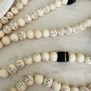 Puede incluir: Un collar de cuentas de madera blancas con caligraf&iacute;a &aacute;rabe negra en cada cuenta. El collar tiene un borla negra con un acento dorado en el extremo.