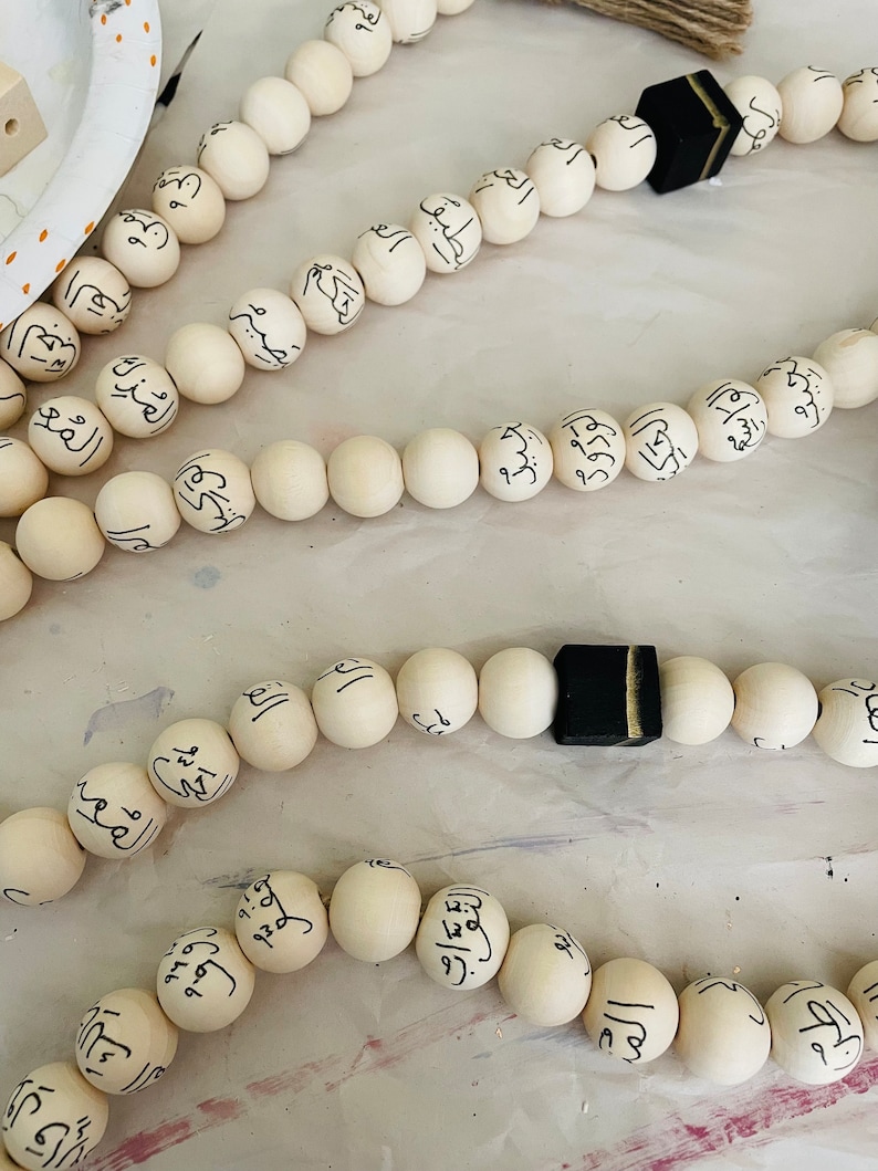 Puede incluir: Un collar de cuentas de madera blancas con caligraf&iacute;a &aacute;rabe negra en cada cuenta. El collar tiene un borla negra con un acento dorado en el extremo.