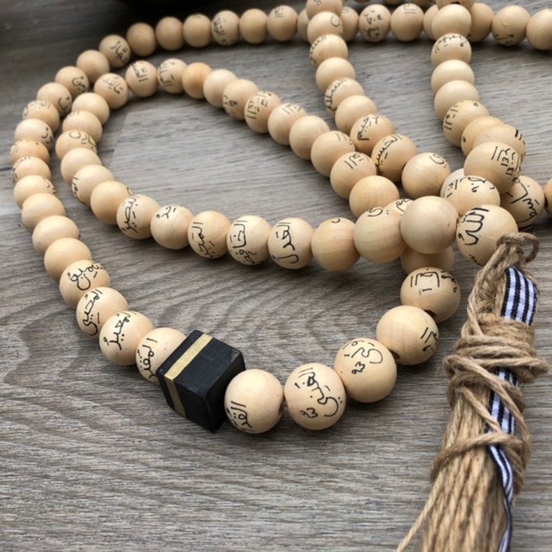 Puede incluir: Un collar de cuentas de oraci&oacute;n de madera con un dije de cubo negro y dorado. Las cuentas est&aacute;n inscritas con caligraf&iacute;a &aacute;rabe. El collar tiene un borla hecha de fibra natural con una cinta a rayas blancas y negras.