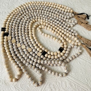 Puede incluir: Una colecci&oacute;n de collares de cuentas de madera blanca con letras negras y borlas. Los collares est&aacute;n dispuestos en un patr&oacute;n circular sobre una superficie blanca.