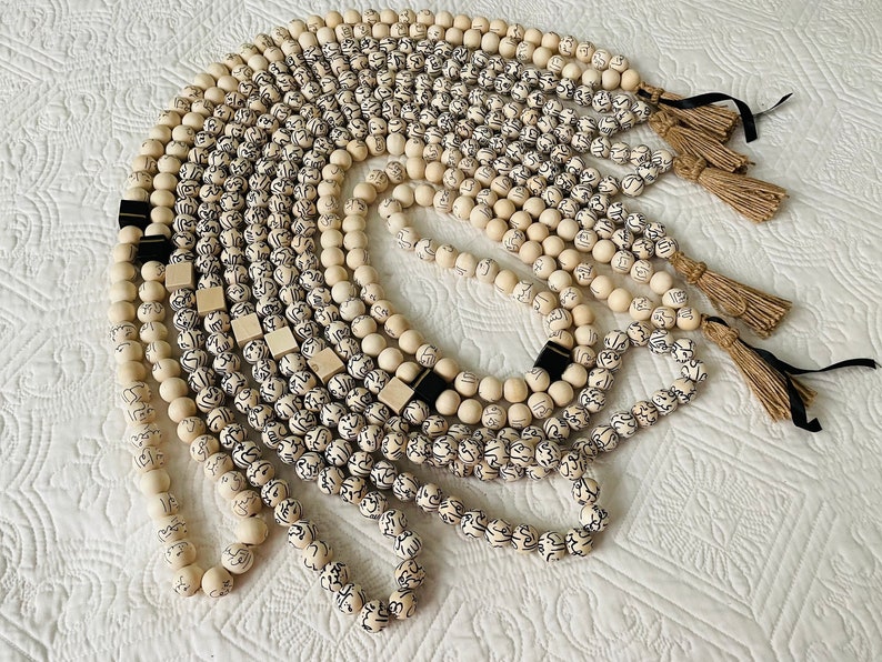 Puede incluir: Una colecci&oacute;n de collares de cuentas de madera blanca con letras negras y borlas. Los collares est&aacute;n dispuestos en un patr&oacute;n circular sobre una superficie blanca.