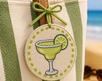 May include: A round, embroidered hanging ornament featuring a cross-stitch design of a light green margarita glass with a lime slice garnish. The ornament is attached to a tan and green striped tote bag with a green fabric bow and cord.