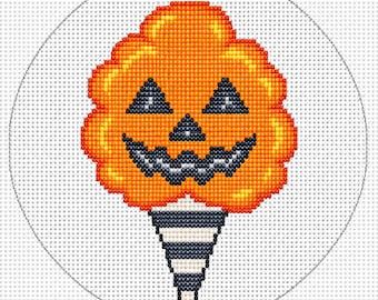 May include: A circular cross-stitch pattern featuring a bright orange pumpkin with a jack-o'-lantern face, including triangular black eyes, a small nose, and a jagged mouth, set atop a black and white striped cone on a white grid background.
