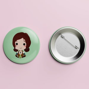Puede incluir: Un botón verde con una ilustración de dibujos animados de una mujer con cabello castaño, vistiendo un chaleco marrón y una camisa verde, sosteniendo una botella de poción verde. La parte posterior del botón es plateada con un alfiler.