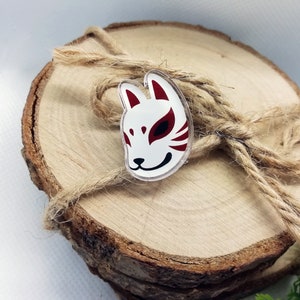 May include: A white and red fox mask pin with black eyes. The pin is on a wooden surface with twine.