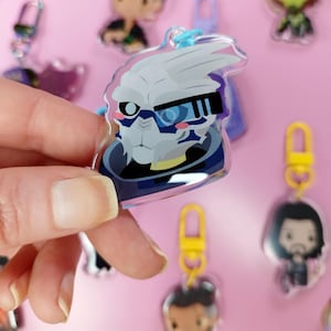 May include: A clear acrylic keychain featuring a stylized character with white and gray skin, blue accents, and a blue and black visor. The keychain has a blue loop and is held against a pink background with other keychains.