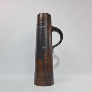 May include: A tall, ceramic pitcher with a handle. The pitcher is primarily brown with dark brown vertical streaks and a black handle. The top portion is a darker shade of brown. The pitcher has a label near the top.
