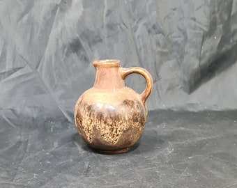 May include: A small, brown ceramic pitcher with a rounded body and a handle. The pitcher has a dark brown base and a lighter brown top. The surface has a mottled, textured appearance.
