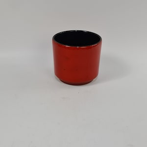 Op de afbeelding: Een rode keramische beker met een zwarte rand. De beker is leeg en staat op een wit oppervlak.