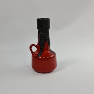 Puede incluir: Un pequeño jarrón de cerámica rojo con un esmalte negro que gotea por el lado. El jarrón tiene una asa y una base redonda.