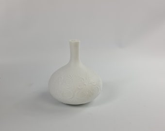 May include: A white ceramic vase with an embossed floral design. The vase has a round body and a narrow neck.