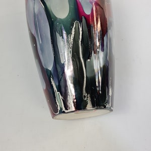 Puede incluir: Un jarr&oacute;n decorativo con un acabado brillante. El jarr&oacute;n presenta una mezcla de colores, incluyendo plata, negro, gris y vetas de rosa y verde. Los colores parecen gotear por los lados. La base del jarr&oacute;n es blanca.