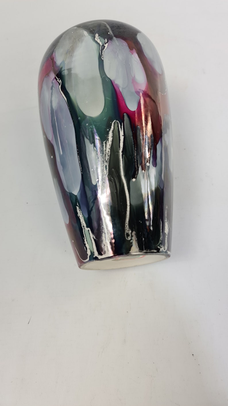 Puede incluir: Un jarr&oacute;n decorativo con un acabado brillante. El jarr&oacute;n presenta una mezcla de colores, incluyendo plata, negro, gris y vetas de rosa y verde. Los colores parecen gotear por los lados. La base del jarr&oacute;n es blanca.