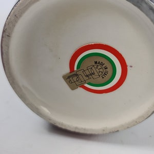 Puede incluir: Un jarr&oacute;n de cer&aacute;mica con interior blanco y exterior oscuro. La base presenta una pegatina con el texto "MADE IN ITALY" en un dise&ntilde;o circular con colores rojo, blanco y verde.