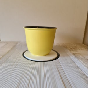 May include: A yellow ceramic planter with a black rim, set on a white coaster with a black border. The planter is centered on a textured, white surface.