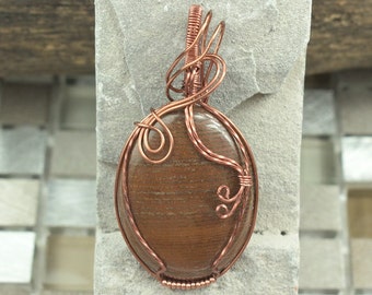 Puede incluir: Un colgante de alambre de cobre envuelto alrededor de una piedra de ágata marrón. El alambre está retorcido y enrollado en un diseño decorativo.