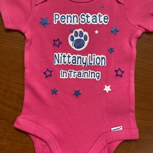 May include: Pink baby onesie with a white paw print and the text "Penn State Nittany Lion in Training" in white with blue glitter stars.