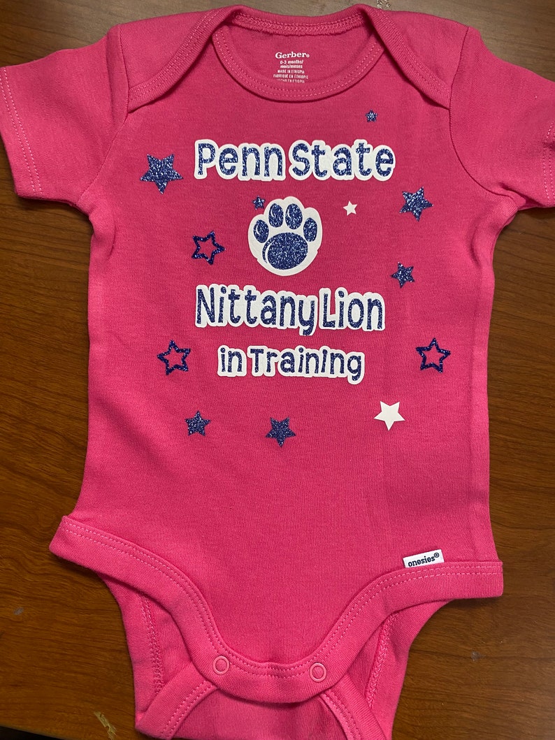 May include: Pink baby onesie with a white paw print and the text "Penn State Nittany Lion in Training" in white with blue glitter stars.