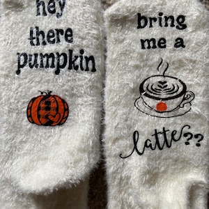 May include: A pair of white fuzzy socks with a pumpkin and the text "hey there pumpkin" on one sock and a coffee cup with the text "bring me a latte?" on the other sock.