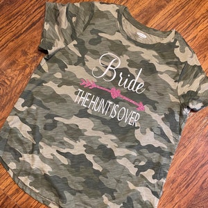 May include: Camouflage-patterned t-shirt with the words "Bride" and "The Hunt Is Over" in white script and a pink glitter arrow design. The shirt is a crew neck style.