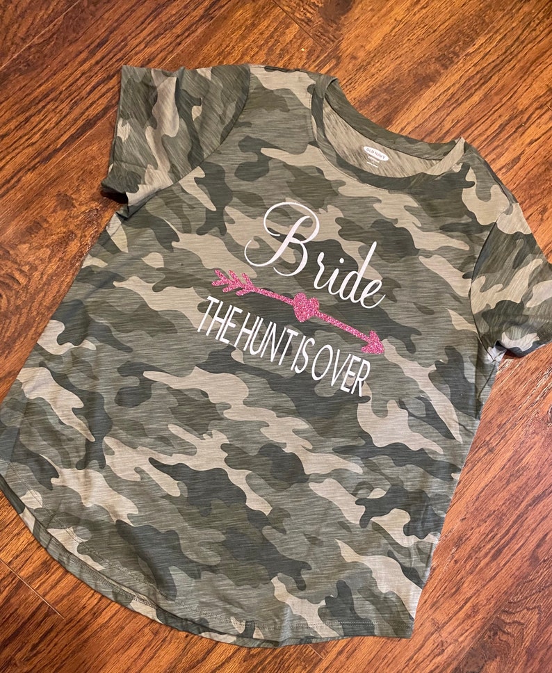 May include: Camouflage-patterned t-shirt with the words "Bride" and "The Hunt Is Over" in white script and a pink glitter arrow design. The shirt is a crew neck style.