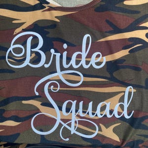 May include: Camouflage-patterned t-shirt with the words "Bride Squad" in a cursive font. The shirt features a mix of brown, green, and black colors, creating a military-inspired design. The text is in a light, silvery color.