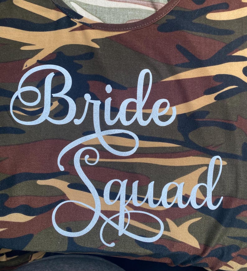 May include: Camouflage-patterned t-shirt with the words "Bride Squad" in a cursive font. The shirt features a mix of brown, green, and black colors, creating a military-inspired design. The text is in a light, silvery color.