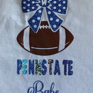 May include: White t-shirt with a blue and white glittery bow, a brown football, and the text "Penn State Babe" in blue glitter.