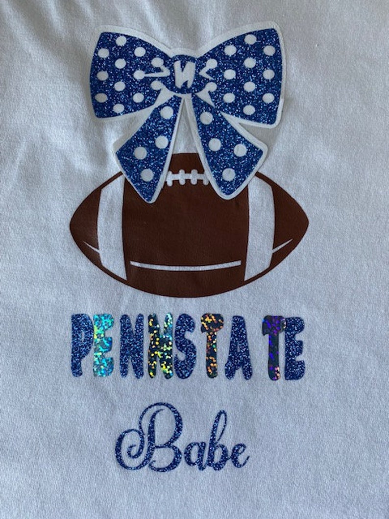 May include: White t-shirt with a blue and white glittery bow, a brown football, and the text "Penn State Babe" in blue glitter.