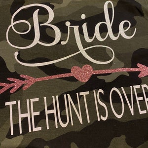 May include: Camouflage-patterned fabric with the word "Bride" in a white, cursive font. Below, a pink glitter arrow points to a pink glitter heart, and the text "THE HUNT IS OVER" is in white block letters.