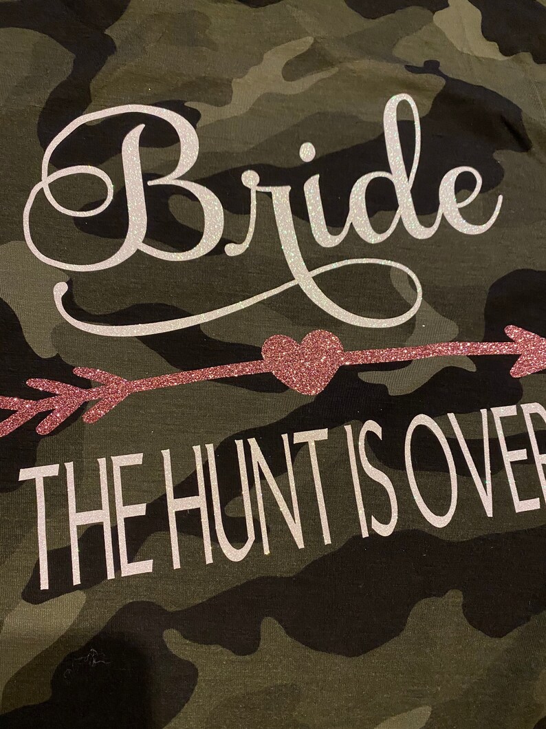 May include: Camouflage-patterned fabric with the word "Bride" in a white, cursive font. Below, a pink glitter arrow points to a pink glitter heart, and the text "THE HUNT IS OVER" is in white block letters.