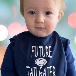 May include: A baby wearing a blue bib with white text that says "FUTURE TAILGATER" and a white outline of a football helmet. The baby is looking at the camera.