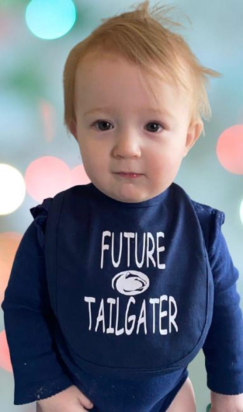 May include: A baby wearing a blue bib with white text that says "FUTURE TAILGATER" and a white outline of a football helmet. The baby is looking at the camera.