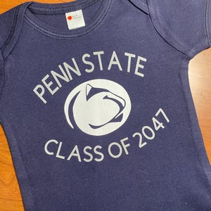 May include: A navy blue baby onesie with a white graphic of the Penn State Nittany Lion mascot and the text "Penn State Class of 2047".