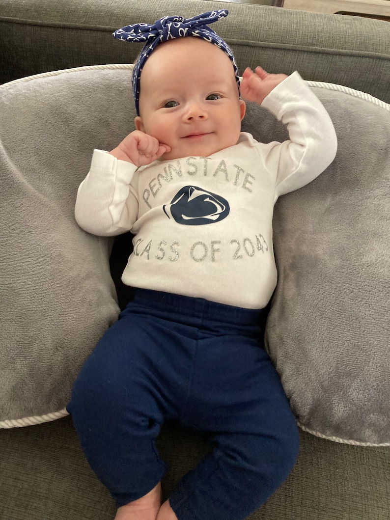 May include: A baby wearing a white onesie with the text "Penn State Class of 2043" and a blue and white bow headband. The baby is wearing blue pants and is sitting on a gray cushion.