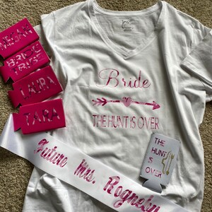 May include: White V-neck t-shirt with "Bride" and "The Hunt Is Over" in pink lettering. Includes pink can coolers with names and "Bride Tribe". A white sash reads "Future Mrs. Rogneby".